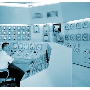 May include: A blue and white control room with a man sitting at a control panel. The panel has many buttons and dials. The man is wearing a white shirt. The room has a sign that says "Happy Father's Day".