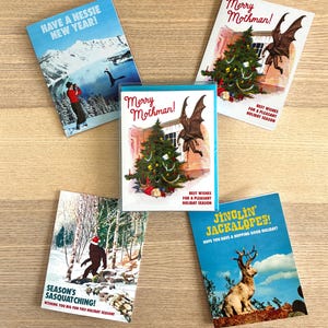 May include: A collection of five holiday greeting cards with vintage-style illustrations. The cards feature cryptids like the Loch Ness Monster, Mothman, Bigfoot, and a Jackalope, with festive text like "Merry Mothman!" and "Season's Sasquatching!"