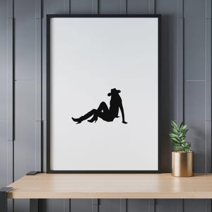 May include: Black silhouette art print of a person wearing a cowboy hat and boots, seated on a white background. The artwork is framed in a black border and displayed on a wooden shelf. A gold pot with a green plant is on the right.