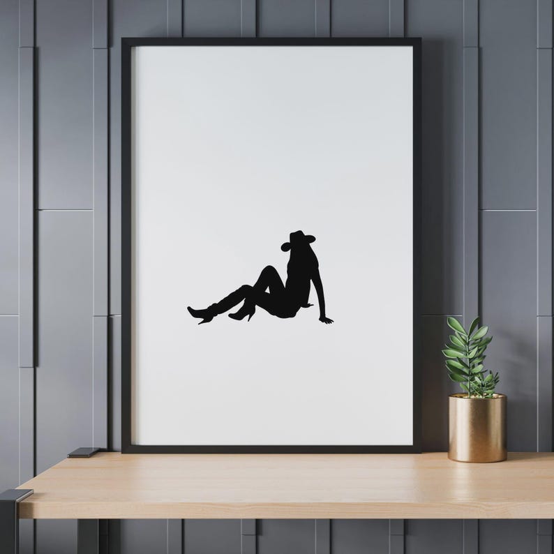 May include: Black silhouette art print of a person wearing a cowboy hat and boots, seated on a white background. The artwork is framed in a black border and displayed on a wooden shelf. A gold pot with a green plant is on the right.