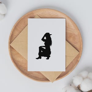 May include: A white card with a black silhouette of a person wearing a cowboy hat, boots, and jeans, sitting on a wooden tray with a brown envelope. The card is centered on the tray, with the envelope partially visible.