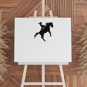 May include: A black silhouette of a person riding a rearing horse, set against a white canvas. The rider wears a hat and is seated on the horse. The image is displayed on a wooden easel, with a textured wooden background.