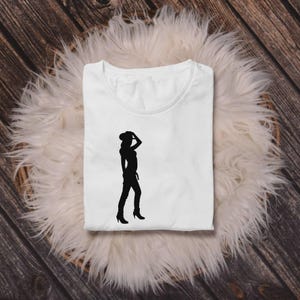 May include: White t-shirt with a black silhouette of a person wearing a cowboy hat, boots, and jeans. The person is posed with one hand on their hat. The shirt is folded on a fluffy white surface.