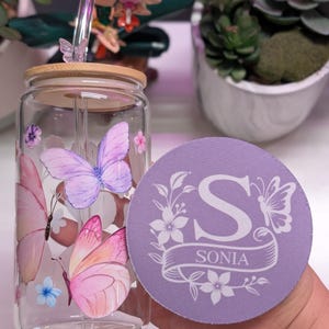May include: A clear glass tumbler with a bamboo lid and a glass straw, decorated with pink and purple butterfly illustrations. A matching lavender coaster with the name "Sonia" and the letter "S" is also visible.