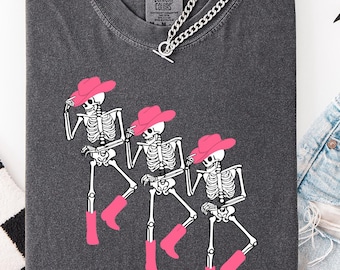 May include: Dark gray Comfort Colors t-shirt featuring a graphic of three dancing skeletons wearing pink cowboy hats and boots. A silver chain necklace rests on the shirt. The shirt is paired with denim shorts and a black and white checkered scarf.