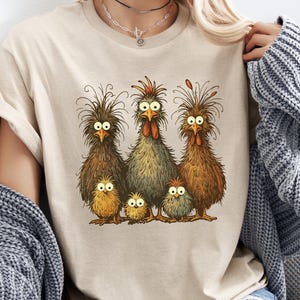 May include: Beige t-shirt featuring a whimsical illustration of a chicken family. The design includes five chickens with expressive eyes and detailed feathering in shades of brown, yellow, and green. The shirt is a casual, everyday wear item.