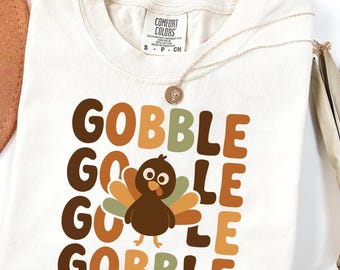 May include: Cream-colored t-shirt with the words "GOBBLE GOBBLE GOBBLE GOBBLE" in brown, green, and orange block letters. A cartoon turkey is centered below the words. A gold necklace and a brown hat are visible.