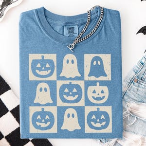 May include: A blue Comfort Colors t-shirt featuring a grid pattern of jack-o'-lanterns and ghosts in a cream color. A silver chain necklace rests on the shirt. The shirt is part of a Halloween theme.