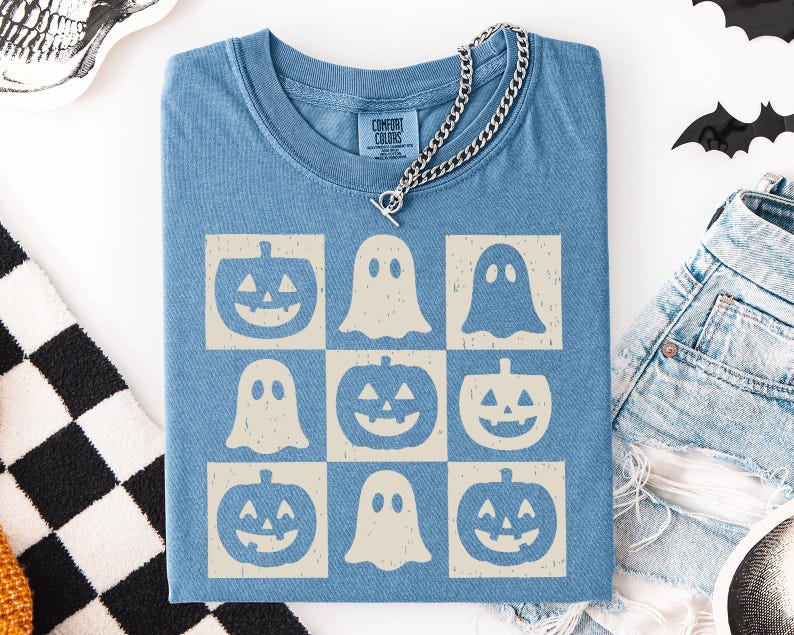 May include: A blue Comfort Colors t-shirt featuring a grid pattern of jack-o'-lanterns and ghosts in a cream color. A silver chain necklace rests on the shirt. The shirt is part of a Halloween theme.