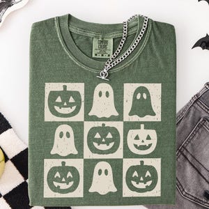 May include: Green t-shirt with a grid pattern of jack-o'-lanterns and ghosts. The shirt has a crew neck and short sleeves. A silver chain necklace with a toggle clasp is draped over the shirt. The shirt is made of a soft, comfortable material.