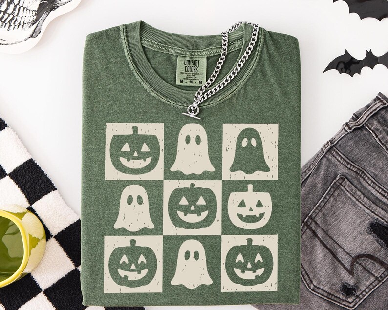 May include: Green t-shirt with a grid pattern of jack-o'-lanterns and ghosts. The shirt has a crew neck and short sleeves. A silver chain necklace with a toggle clasp is draped over the shirt. The shirt is made of a soft, comfortable material.