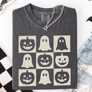 May include: Dark gray t-shirt featuring a grid of Halloween-themed graphics. The design includes jack-o'-lanterns and ghost figures in a cream color. A silver chain necklace is draped over the shirt. The shirt is folded and placed on a white surface.