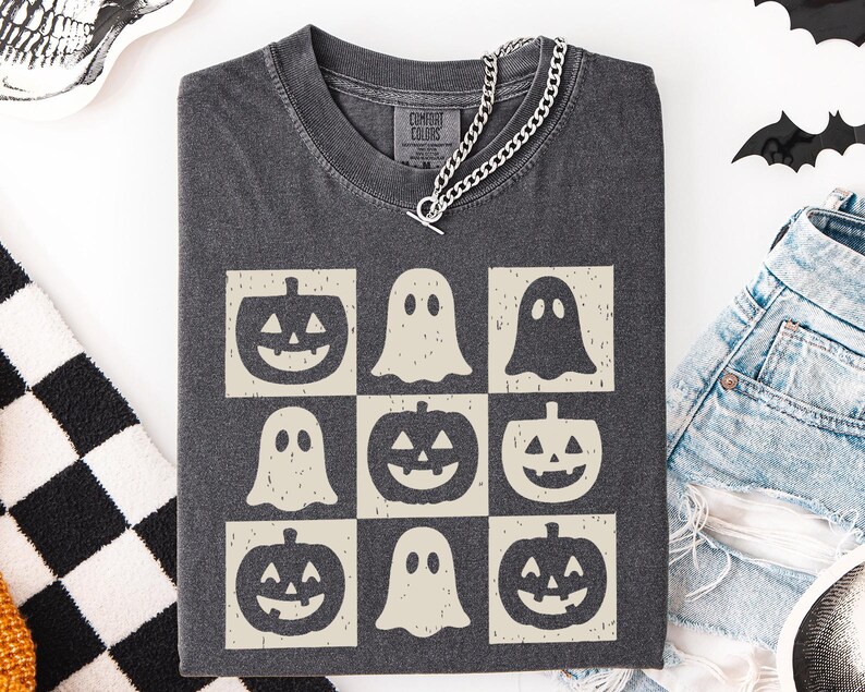 May include: Dark gray t-shirt featuring a grid of Halloween-themed graphics. The design includes jack-o'-lanterns and ghost figures in a cream color. A silver chain necklace is draped over the shirt. The shirt is folded and placed on a white surface.