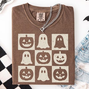 May include: Brown t-shirt with a grid pattern of jack-o'-lanterns and ghosts. The shirt has a crew neck and short sleeves. A silver chain necklace is draped over the shirt. The shirt is folded on a white surface.