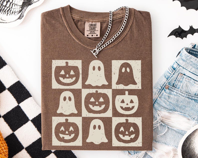 May include: Brown t-shirt with a grid pattern of jack-o'-lanterns and ghosts. The shirt has a crew neck and short sleeves. A silver chain necklace is draped over the shirt. The shirt is folded on a white surface.