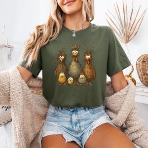 May include: Olive green t-shirt featuring a whimsical illustration of a chicken family with wide-eyed expressions. The graphic includes three adult chickens and three chicks. The shirt is a comfortable, casual style.