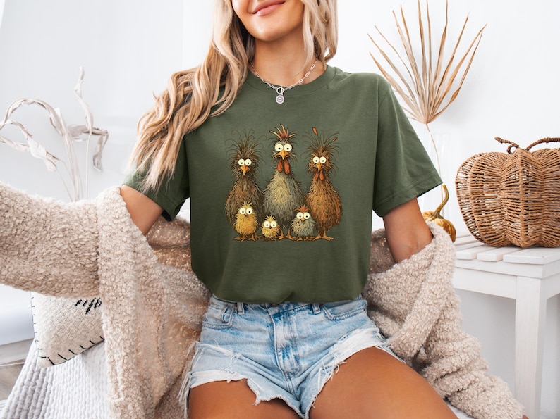 May include: Olive green t-shirt featuring a whimsical illustration of a chicken family with wide-eyed expressions. The graphic includes three adult chickens and three chicks. The shirt is a comfortable, casual style.