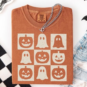 May include: A rust-colored t-shirt featuring a grid pattern of jack-o'-lanterns and ghosts. A silver chain necklace with a toggle clasp is draped across the shirt. The shirt is folded, and a pair of denim shorts are visible.