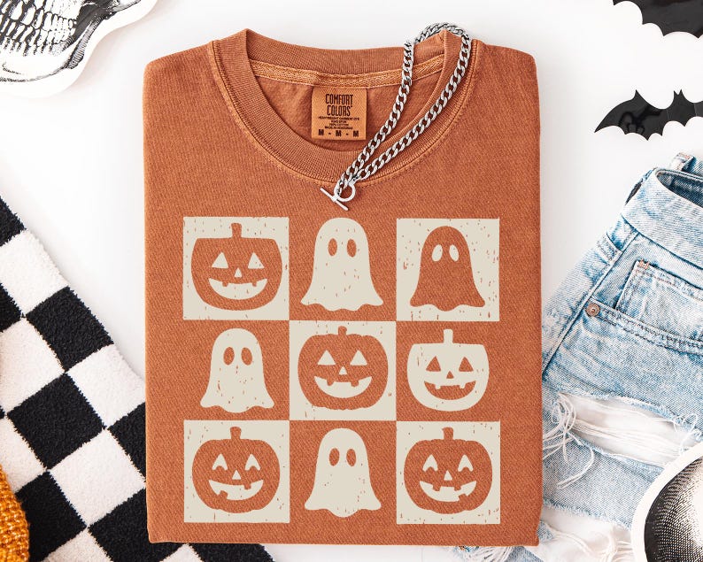 May include: A rust-colored t-shirt featuring a grid pattern of jack-o'-lanterns and ghosts. A silver chain necklace with a toggle clasp is draped across the shirt. The shirt is folded, and a pair of denim shorts are visible.
