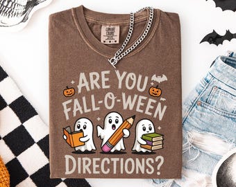 May include: Brown t-shirt with the text "ARE YOU FALL-O-WEEN DIRECTIONS?" and illustrations of ghosts, pumpkins, and a bat. A silver chain necklace is draped across the shirt. The shirt is folded on a white surface.