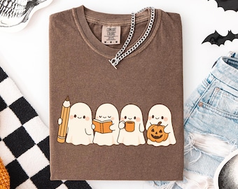 May include: Brown Comfort Colors t-shirt featuring a graphic of four cartoon ghosts, each holding a pencil, book, mug, and pumpkin. A silver chain necklace rests on the shirt. The shirt is folded with a pair of denim shorts and a black and white checkered pattern.