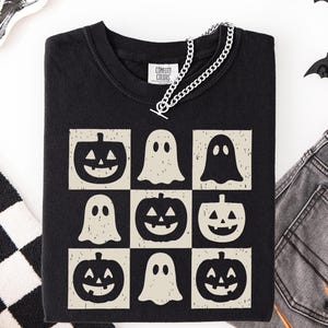 May include: Black Comfort Colors t-shirt featuring a grid design with jack-o'-lanterns and ghost graphics. A silver chain necklace is draped across the neckline. The shirt is a Halloween-themed apparel item.