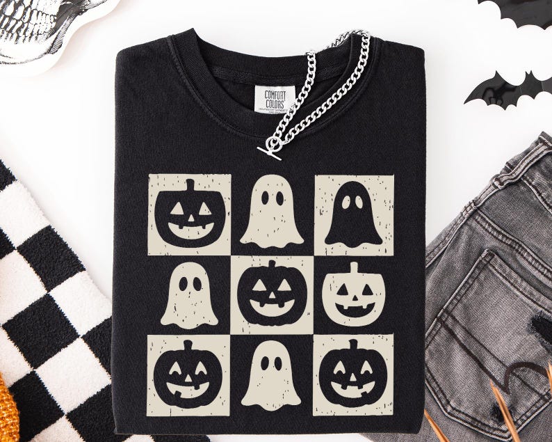May include: Black Comfort Colors t-shirt featuring a grid design with jack-o'-lanterns and ghost graphics. A silver chain necklace is draped across the neckline. The shirt is a Halloween-themed apparel item.