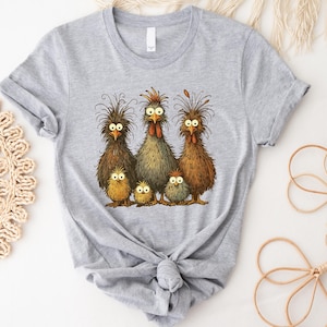 May include: A light gray t-shirt featuring a whimsical illustration of a chicken family. The design includes five chickens with expressive eyes and detailed feather textures in shades of brown, yellow, and gray. The shirt is tied at the waist.