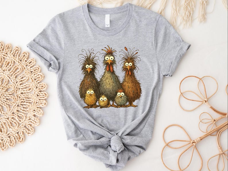 May include: A light gray t-shirt featuring a whimsical illustration of a chicken family. The design includes five chickens with expressive eyes and detailed feather textures in shades of brown, yellow, and gray. The shirt is tied at the waist.