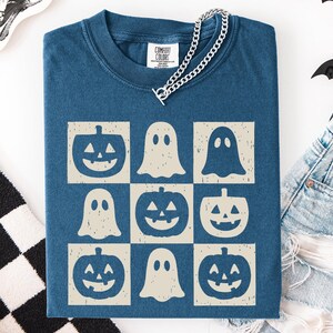 May include: A blue Comfort Colors t-shirt featuring a grid pattern of Halloween-themed graphics. The design includes jack-o'-lanterns and ghost figures in a cream color. A silver chain necklace is draped over the shirt.