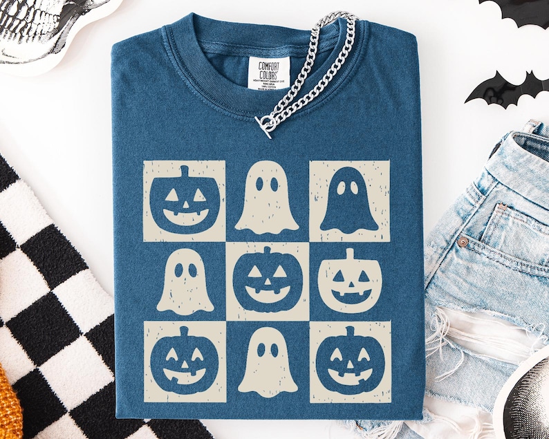 May include: A blue Comfort Colors t-shirt featuring a grid pattern of Halloween-themed graphics. The design includes jack-o'-lanterns and ghost figures in a cream color. A silver chain necklace is draped over the shirt.