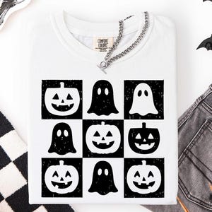 May include: White t-shirt with a grid pattern of black and white Halloween-themed graphics. The design includes jack-o'-lanterns and ghost figures. A silver chain necklace is draped across the neckline. The shirt is folded.