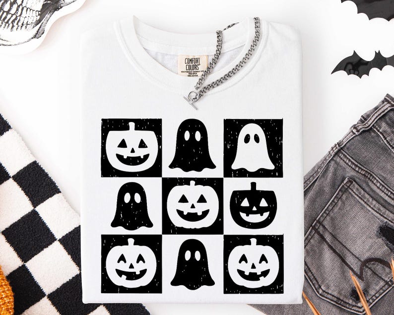 May include: White t-shirt with a grid pattern of black and white Halloween-themed graphics. The design includes jack-o'-lanterns and ghost figures. A silver chain necklace is draped across the neckline. The shirt is folded.