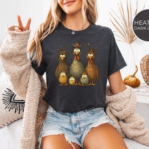 May include: Dark heather gray t-shirt featuring a whimsical illustration of a chicken family. The design includes six chickens with expressive eyes and detailed feathering. The shirt is a comfortable crew neck style.