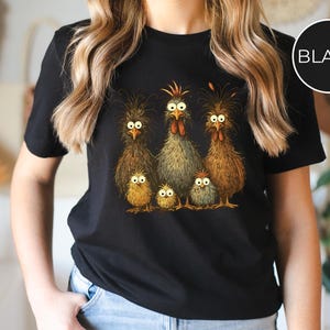May include: Black t-shirt featuring a whimsical illustration of a chicken family. The design includes a rooster, hens, and chicks with expressive eyes and detailed feathering. A fun and unique graphic tee.
