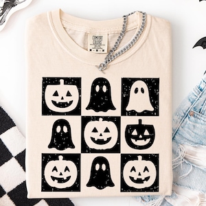 May include: Cream-colored t-shirt featuring a grid of black and white Halloween-themed graphics. The design includes jack-o'-lanterns and ghost figures. The shirt has a crew neck and short sleeves. A silver chain necklace is draped across the shirt.