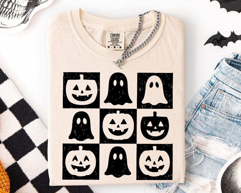 May include: Cream-colored t-shirt featuring a grid of black and white Halloween-themed graphics. The design includes jack-o'-lanterns and ghost figures. The shirt has a crew neck and short sleeves. A silver chain necklace is draped across the shirt.