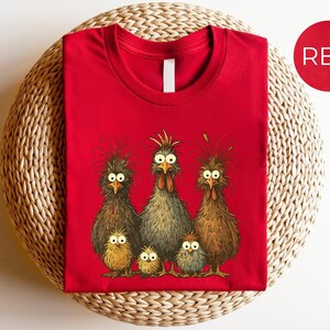 May include: A red t-shirt featuring a whimsical illustration of a chicken family. The design includes three adult chickens and three chicks, all with expressive eyes and detailed feathering. The word "RED" is in a red circle.