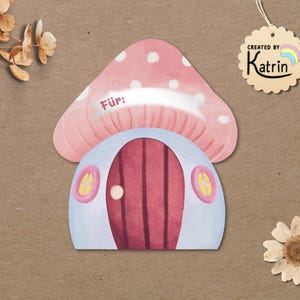 May include: A mushroom-shaped gift tag with a pink cap featuring white spots and a light blue base. The tag has a dark red door, two small yellow windows, and the word "F&uuml;r:" printed on the cap. A scalloped tag reads "CREATED BY Katrin".