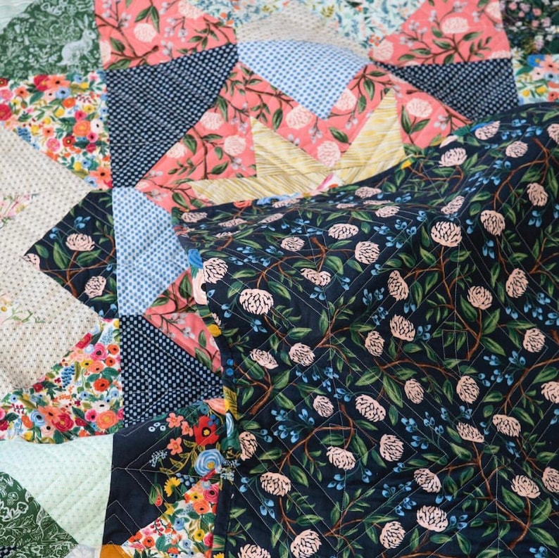 May include: A quilt made of various floral patterned fabrics in shades of pink, blue, green, and white. The quilt is folded in half, revealing a dark blue fabric with a floral pattern in shades of pink and green.