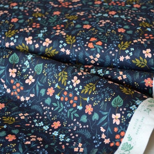 May include: Dark navy fabric with a floral pattern featuring pink, peach, and blue flowers, along with green leaves and stems. The fabric is from the "Wildwood Collection" and has a white selvage edge.