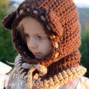 May include: A brown crocheted hooded scarf with a beige trim. The hood has a pointed tip and the scarf has a tie at the front. The scarf is worn by a child.