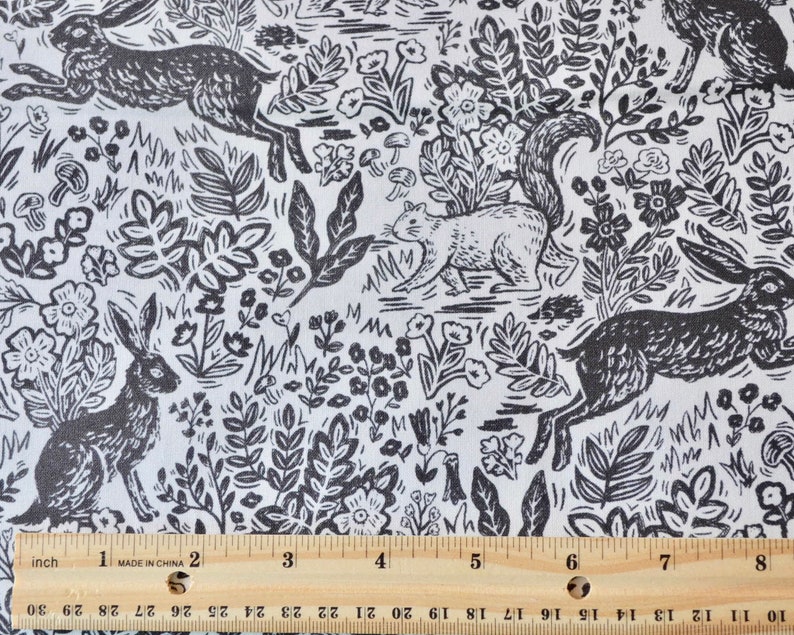 K&ouml;nnte beinhalten: Schwarz-wei&szlig;er Stoff mit einem Waldmuster, das Hasen, ein Eichh&ouml;rnchen, Blumen und Bl&auml;tter zeigt. Das Muster wiederholt sich. Ein Zoll-Lineal ist am unteren Rand zu sehen.