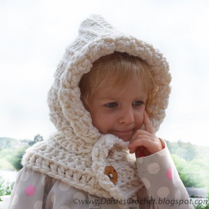 May include: A white crocheted hooded scarf with a button closure. The scarf is worn by a young child with blonde hair.