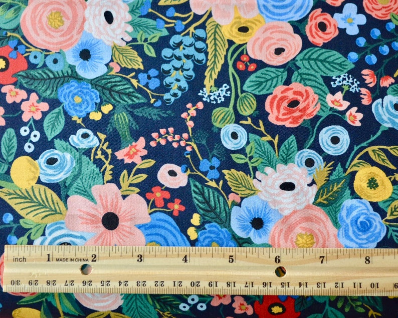 Puede incluir: Tela floral con fondo azul marino. La tela presenta un denso estampado de flores en tonos rosa, azul, amarillo y verde. Una regla de madera con medidas en pulgadas est&aacute; colocada sobre la tela.