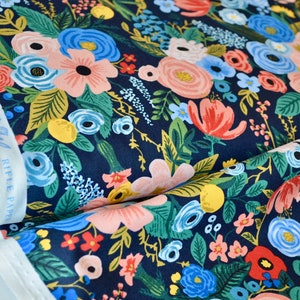 Puede incluir: Tela floral con fondo azul marino. La tela presenta un denso estampado de flores en tonos rosa, azul, rojo y amarillo, con hojas verdes. A la izquierda se ve un rollo de tela blanca con estampado floral azul.