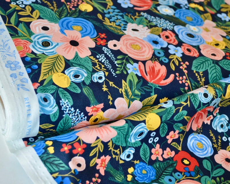 Puede incluir: Tela floral con fondo azul marino. La tela presenta un denso estampado de flores en tonos rosa, azul, rojo y amarillo, con hojas verdes. A la izquierda se ve un rollo de tela blanca con estampado floral azul.