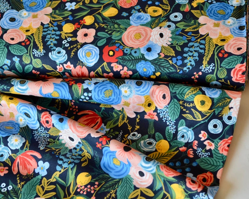 Puede incluir: Tela azul marino oscuro con un denso estampado floral. El dise&ntilde;o presenta una variedad de flores en tonos rosa, azul, amarillo y rojo, sobre un fondo de follaje verde. La tela est&aacute; doblada, mostrando la repetici&oacute;n del patr&oacute;n.