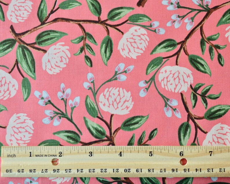May include: Pink fabric with a white floral pattern. The fabric features large white flowers with green leaves and smaller white flowers with light blue centers. A ruler is placed at the bottom of the image for scale.