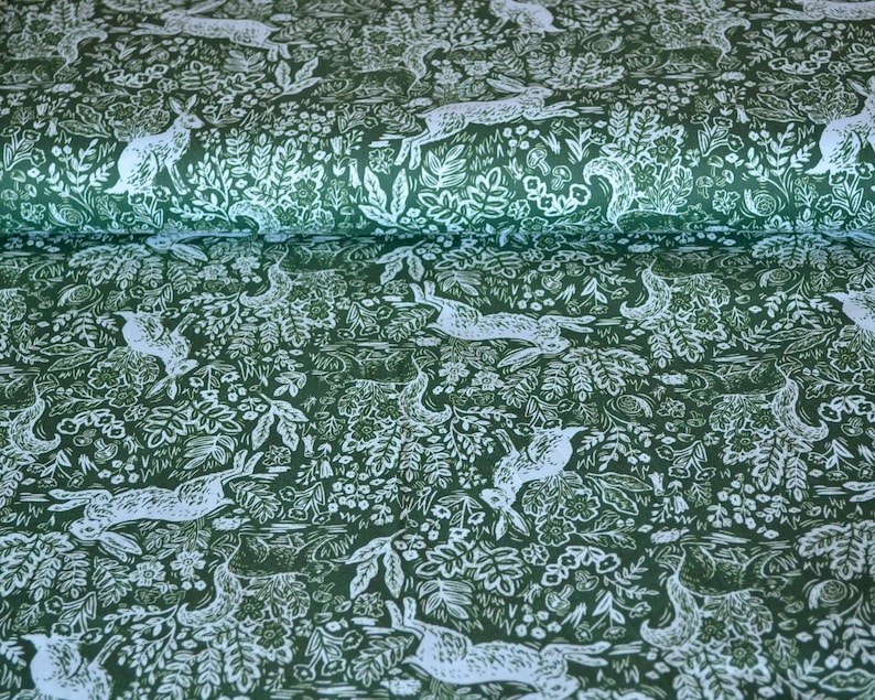 Puede incluir: Tela verde oscuro con un patr&oacute;n repetido de liebres blancas y elementos bot&aacute;nicos. El dise&ntilde;o presenta ilustraciones detalladas de conejos entre hojas, flores y otros elementos naturales, creando un tema de bosque.